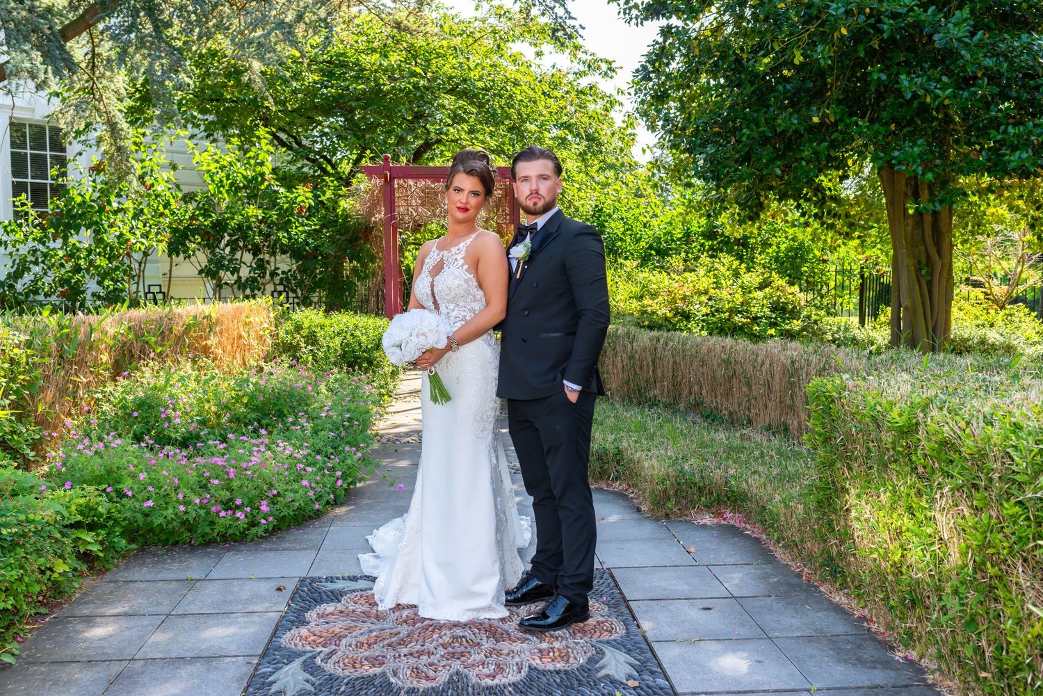 Bride and groom posing on a garden pathway surrounded by lush greenery on their wedding day.