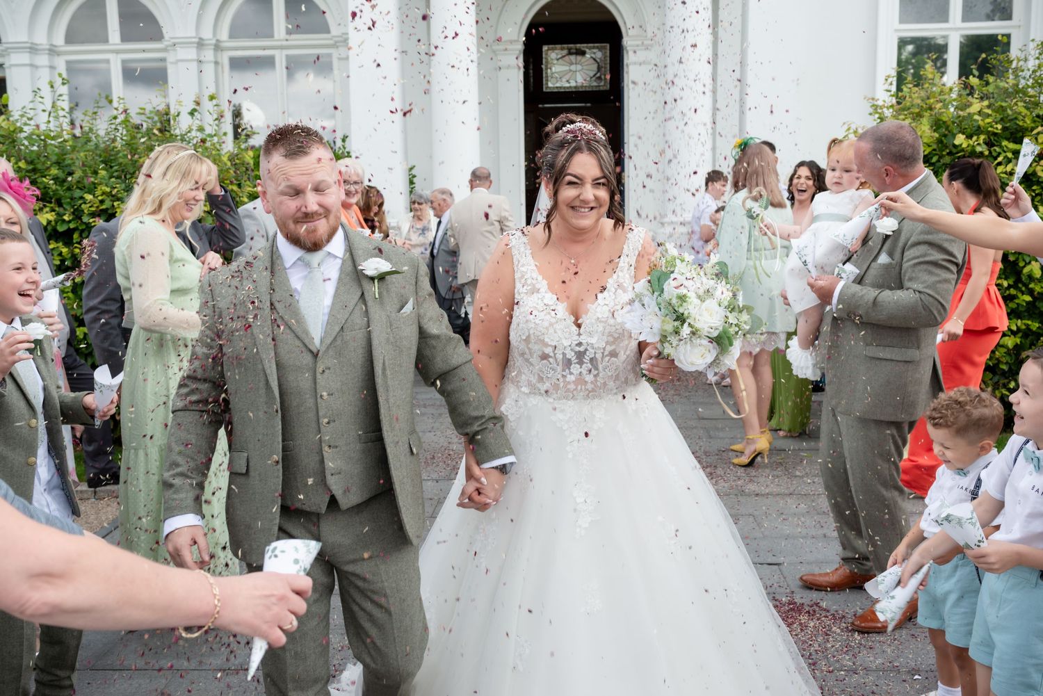 Newlywed couple walks through confetti shower outside white building, bride in ballgown holding bouquet, groom in sage tweed suit.