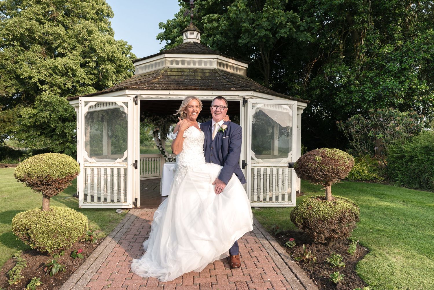 Bride and groom posing in front of a white gazebo in a manicured garden on their wedding day.