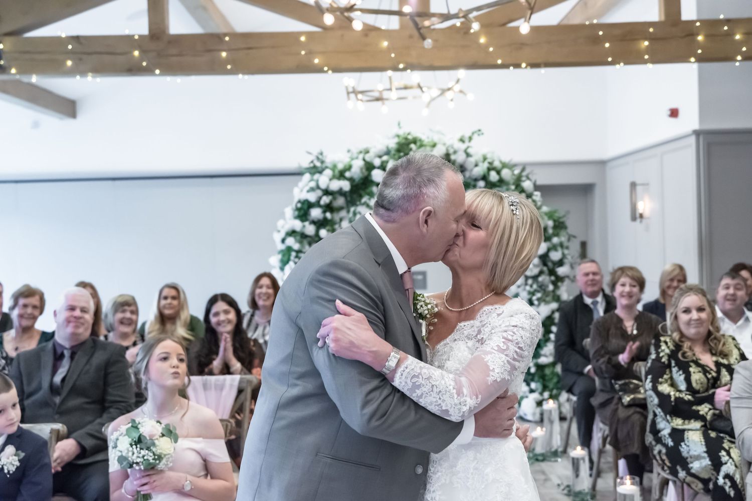 Bride and groom share first kiss at wedding ceremony with floral arch and seated guests in elegant venue.