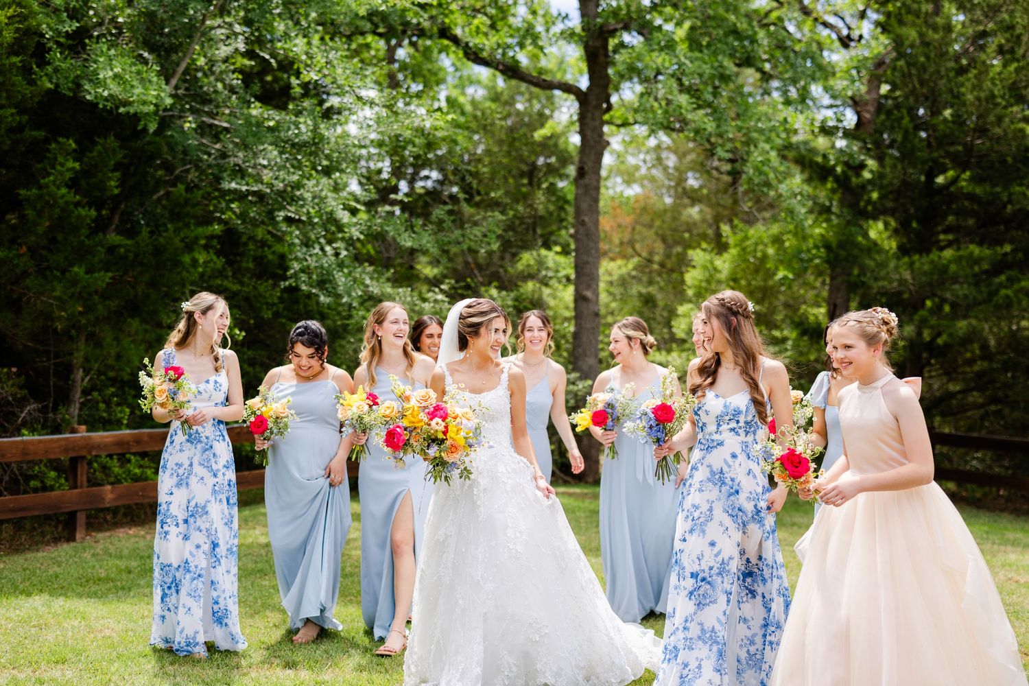 A bridal party in blue floral and light blue dresses poses outdoors with colorful bouquets.