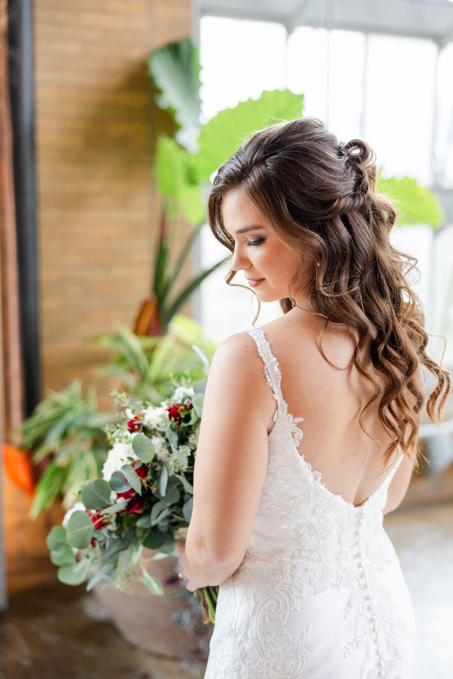 A bride in a lace dress holds a bouquet while posing near indoor plants.