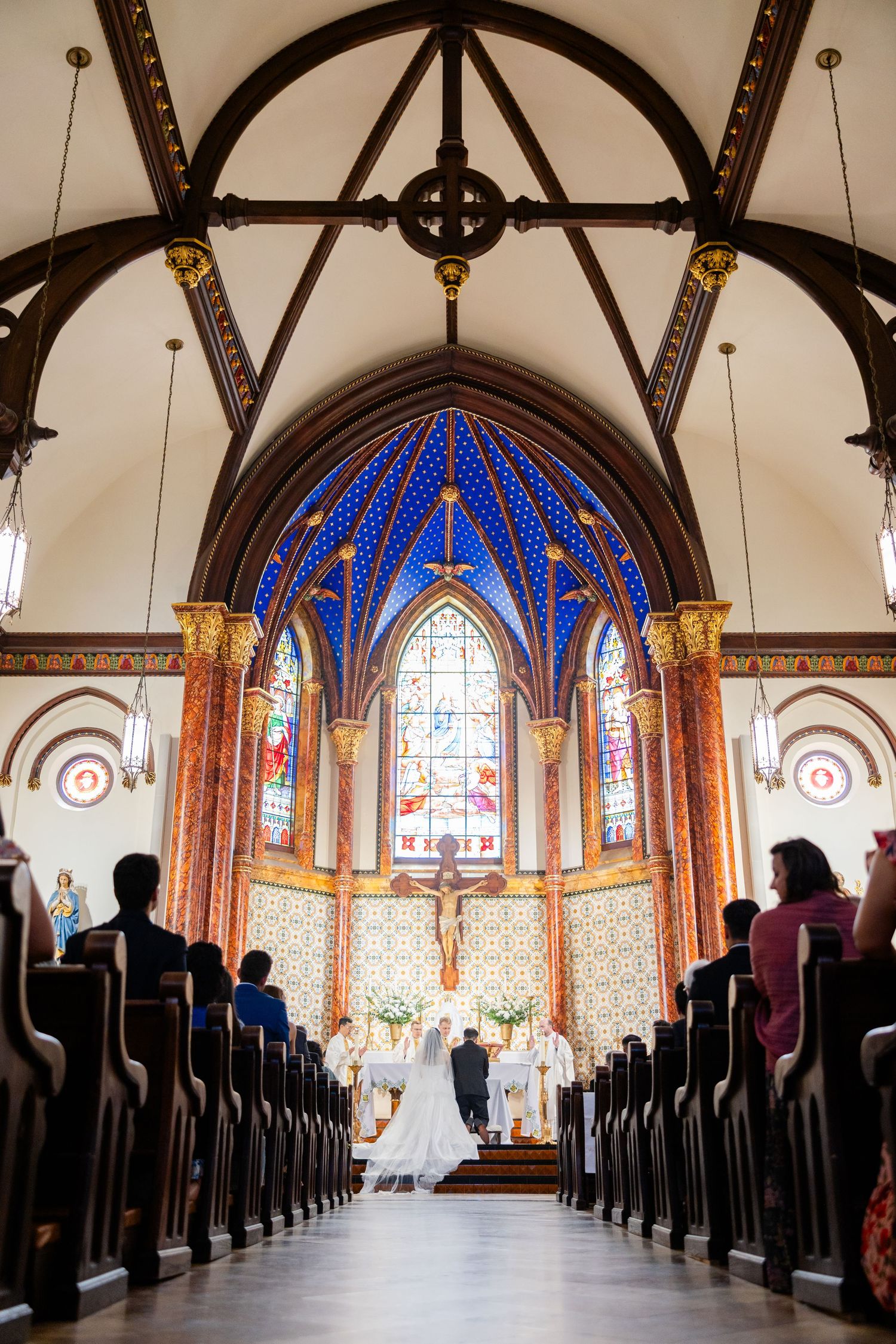 A wedding ceremony takes place in a stunning Gothic chapel with vibrant blue stained glass windows and vaulted wooden beams.