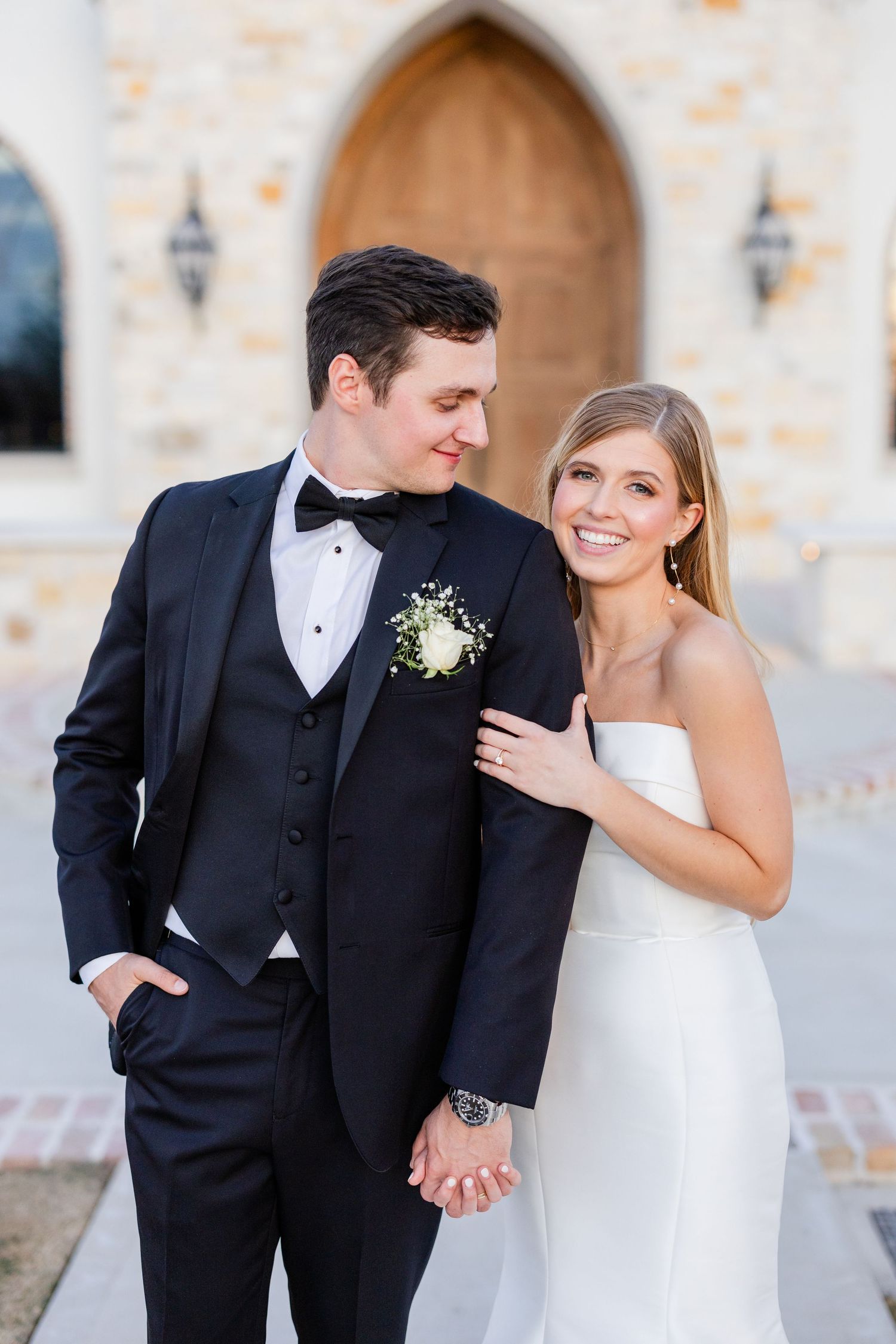 A wedding couple poses together outside a stone chapel in formal attire.