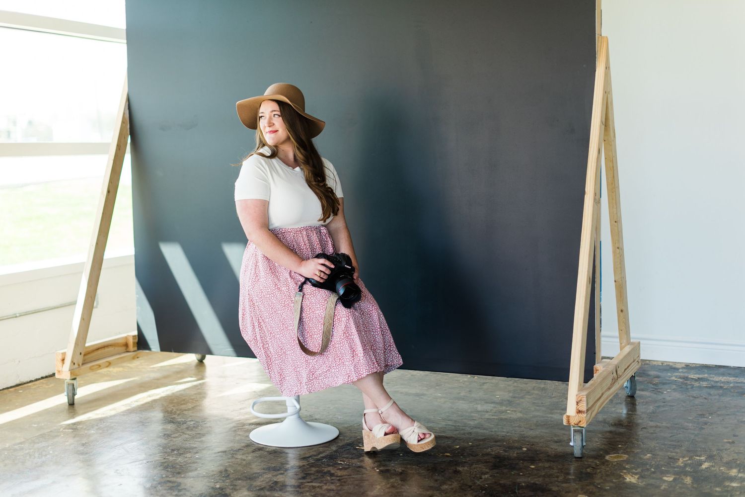 A photographer in a pink skirt and hat poses stylishly on a chair against a dark wall.