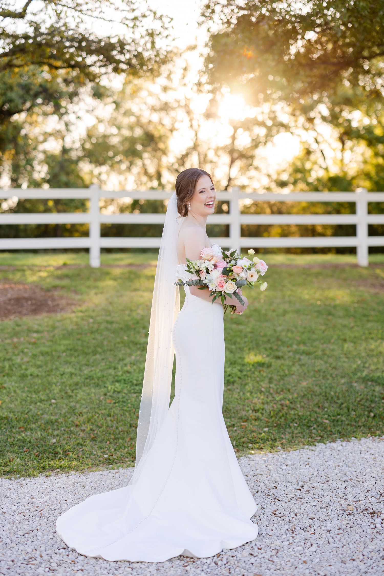 A bride in a flowing white dress holds a bouquet while standing beside a white fence at sunset.