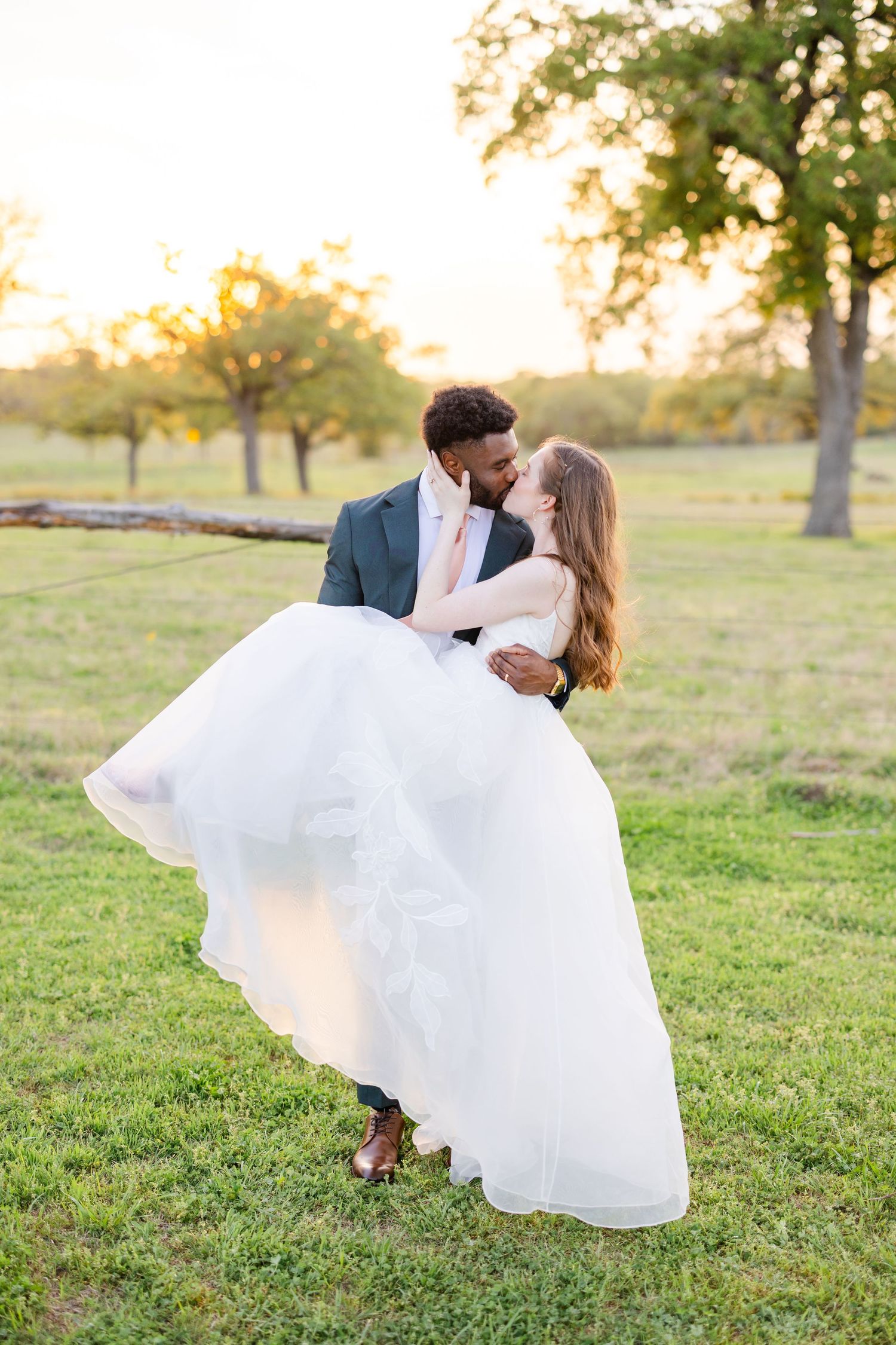 Romantic sunset wedding photo of a couple embracing in a grassy field with golden light filtering through trees.