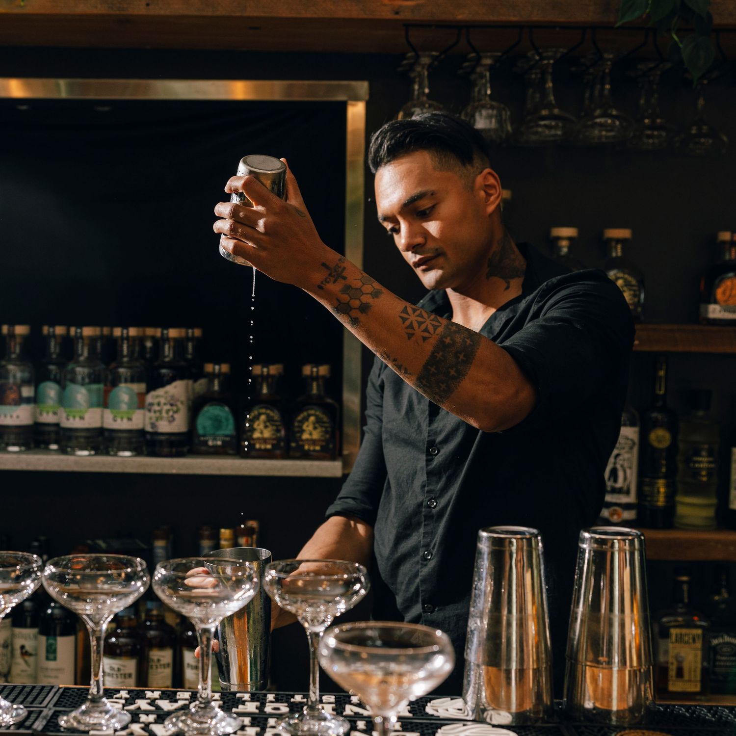 Professional bartender preparing cocktails behind a dark bar counter with backlit bottles.