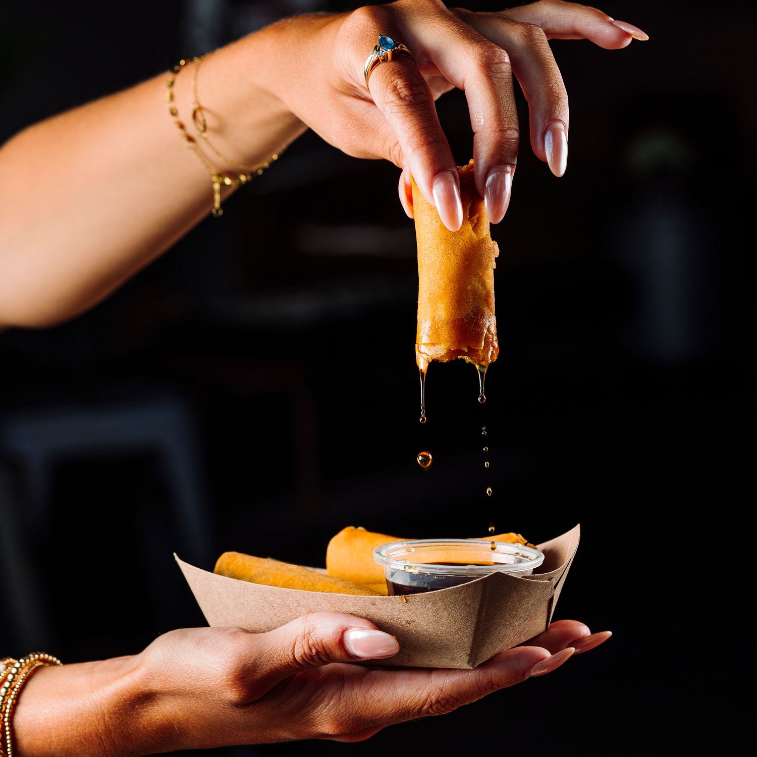 Hands pouring golden sauce over food in paper container.