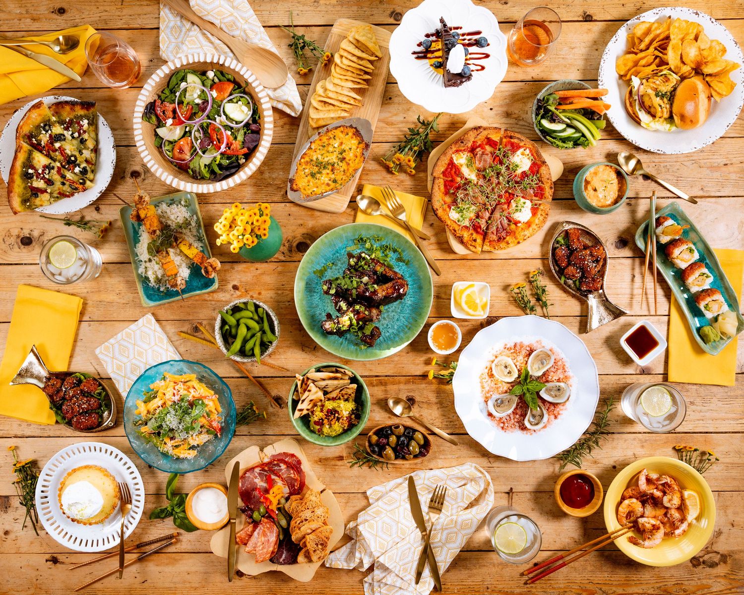 Overhead view of diverse restaurant dishes on wooden table including pizza, salads and appetizers.