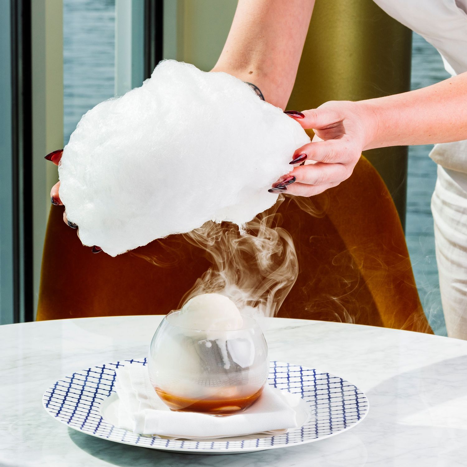 Hands lifting smoke cloche to reveal plated dish on white tablecloth.