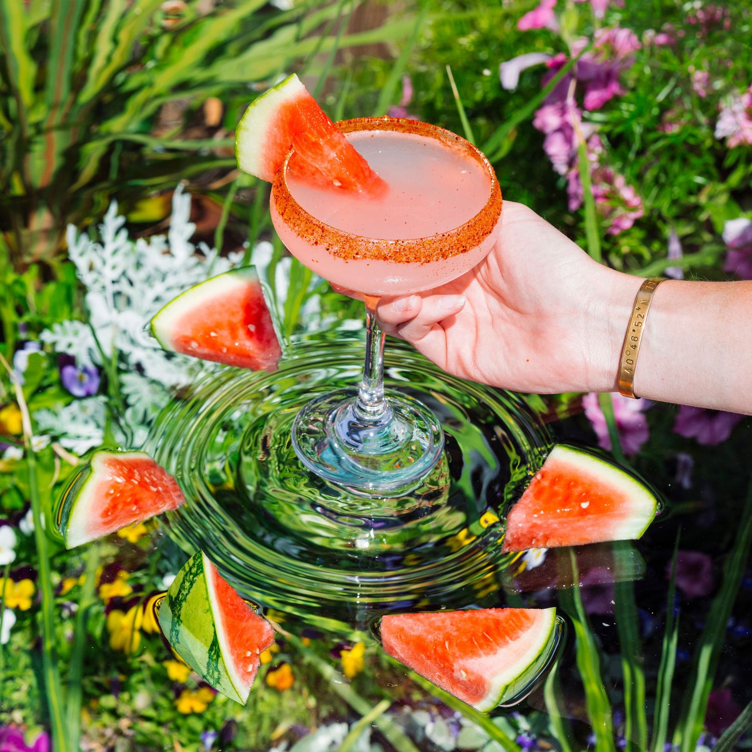 Watermelon-garnished cocktail held over rippling water in garden setting.