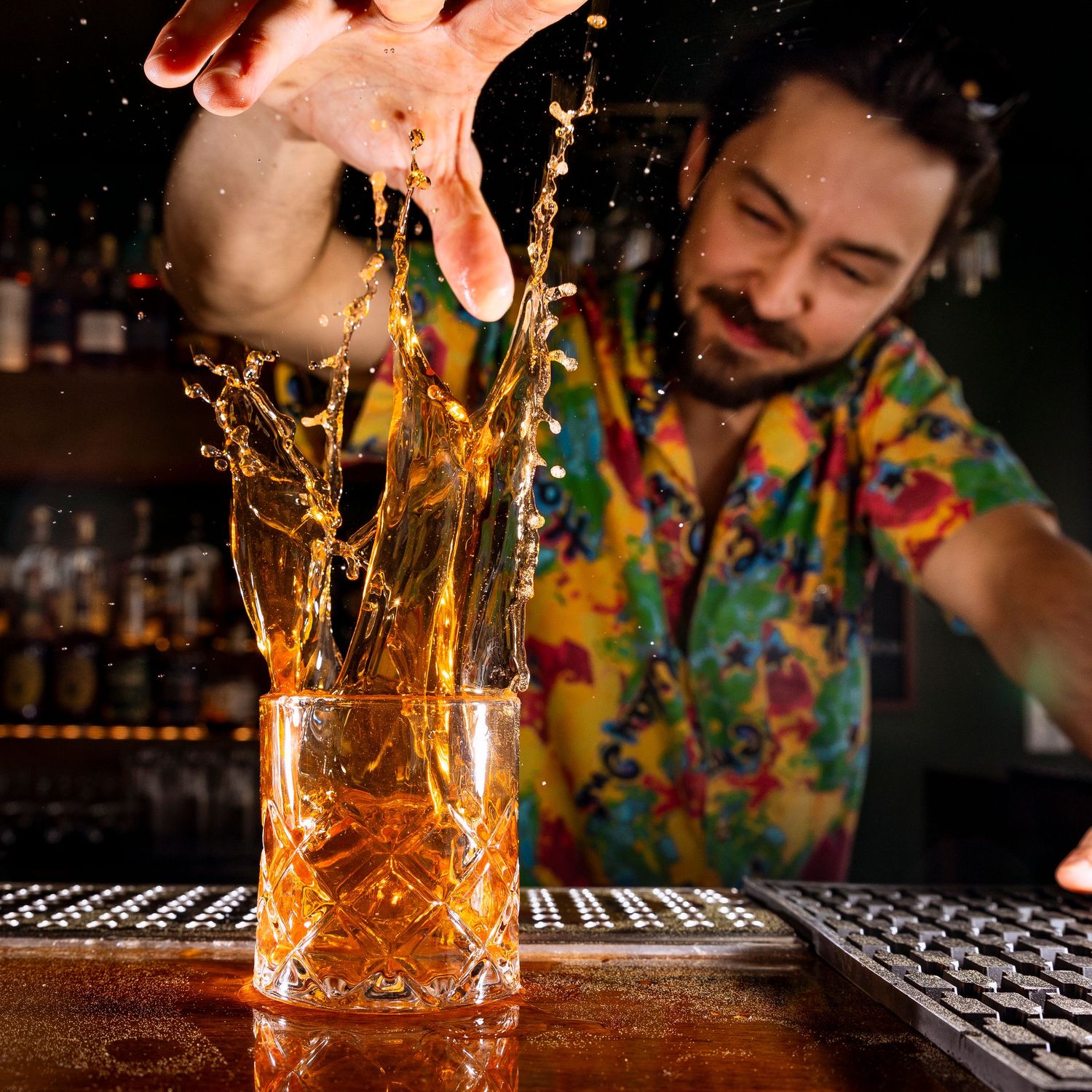 Bartender in a colorful Hawaiian shirt pouring a drink that splashes dramatically in a glass at a bar counter.