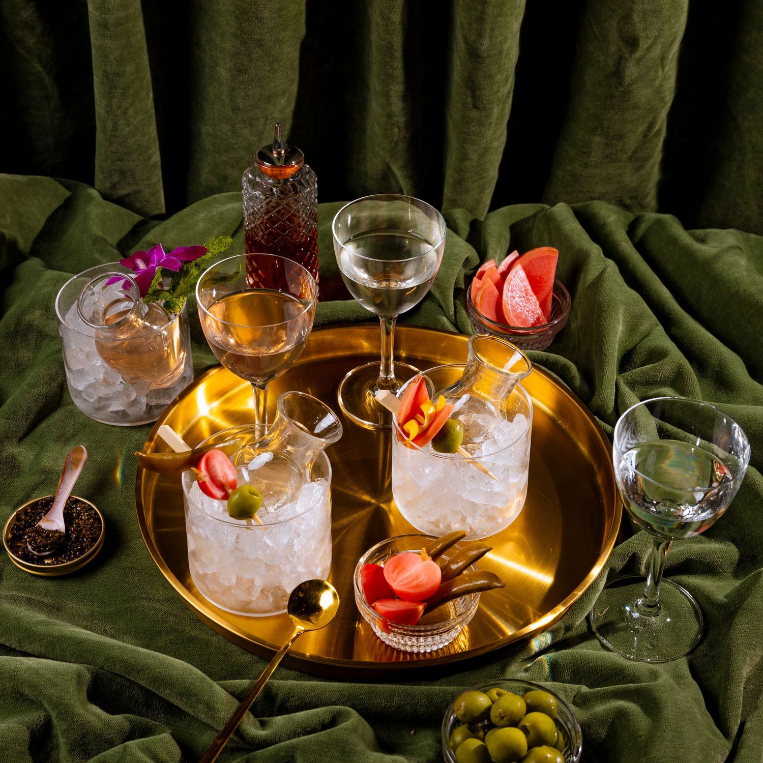 Elegant cocktail spread with garnishes and olives on a green velvet surface with ambient lighting.