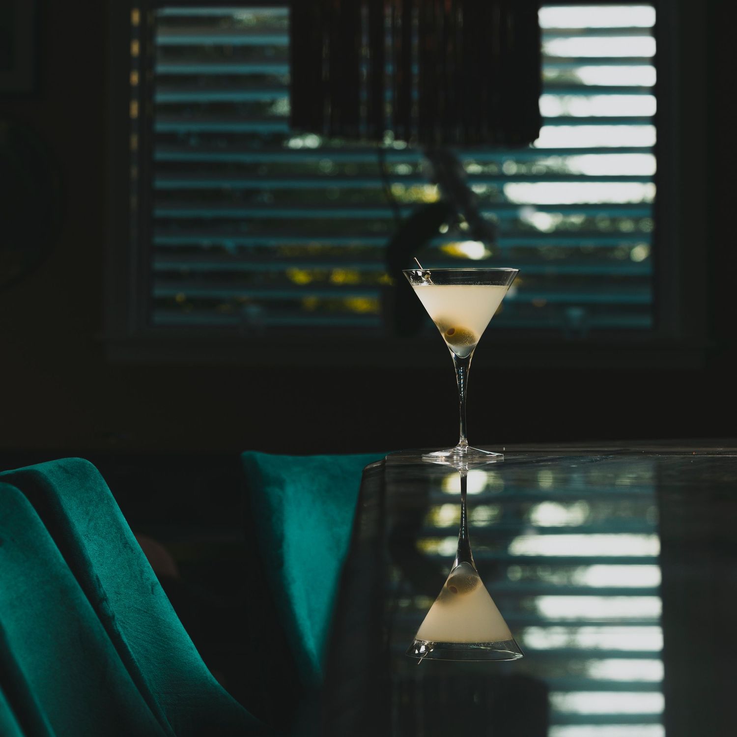 Martini glass reflecting on glossy table surface with venetian blinds in background.
