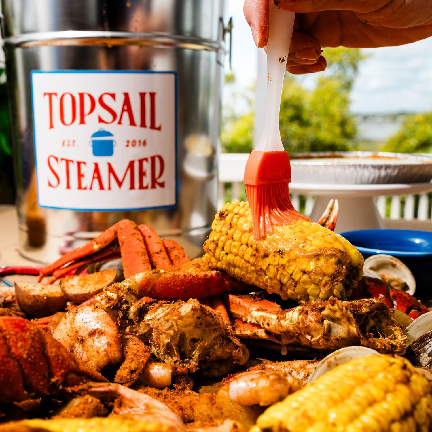 A seafood boil with crab legs, corn, and shellfish next to a Topsail Steamer container.