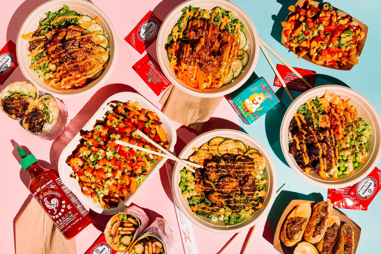 Overhead view of colorful Asian dishes with sriracha sauce bottles on pink and blue background.