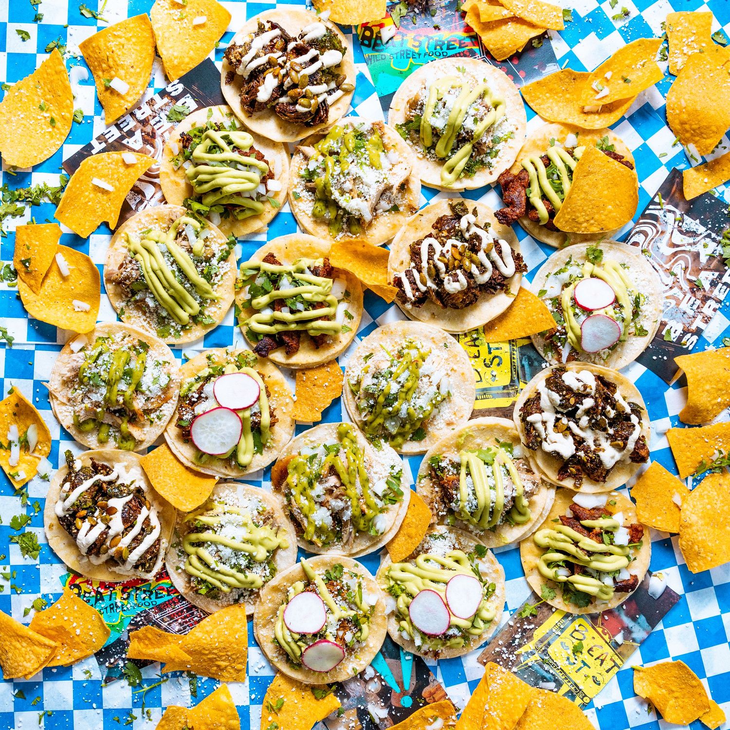 Overhead view of street tacos garnished with fresh vegetables and sauces on blue checkered background.