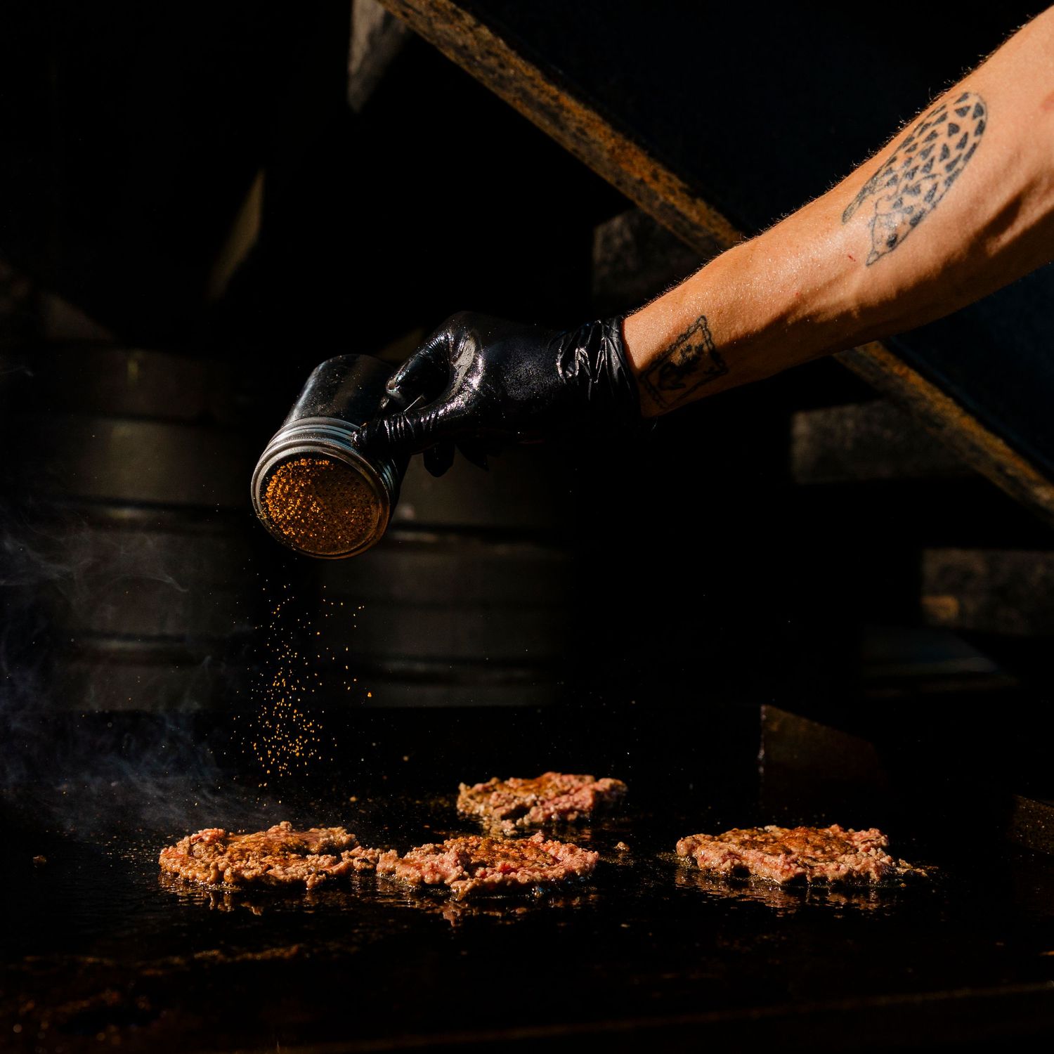 Seasoning being sprinkled onto meat on a dark grill surface.