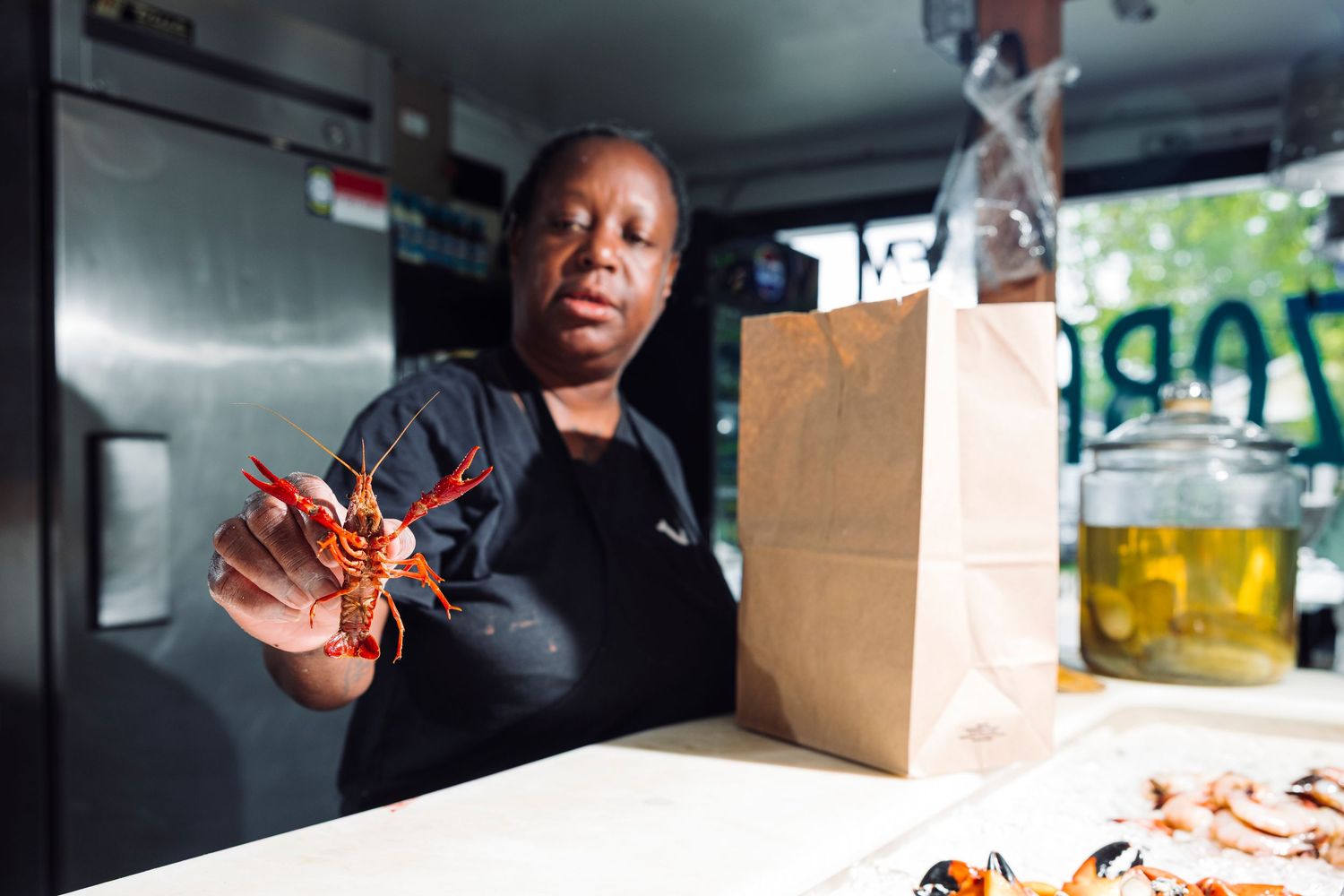 A food service worker in black attire gestures while working inside a food truck with takeout bags visible.