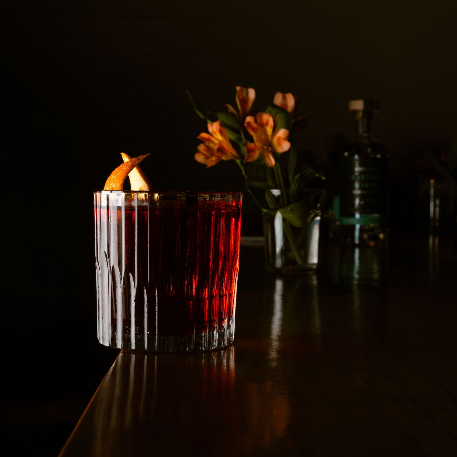 Illuminated bar counter with cocktail and bottles silhouetted in dark ambiance.