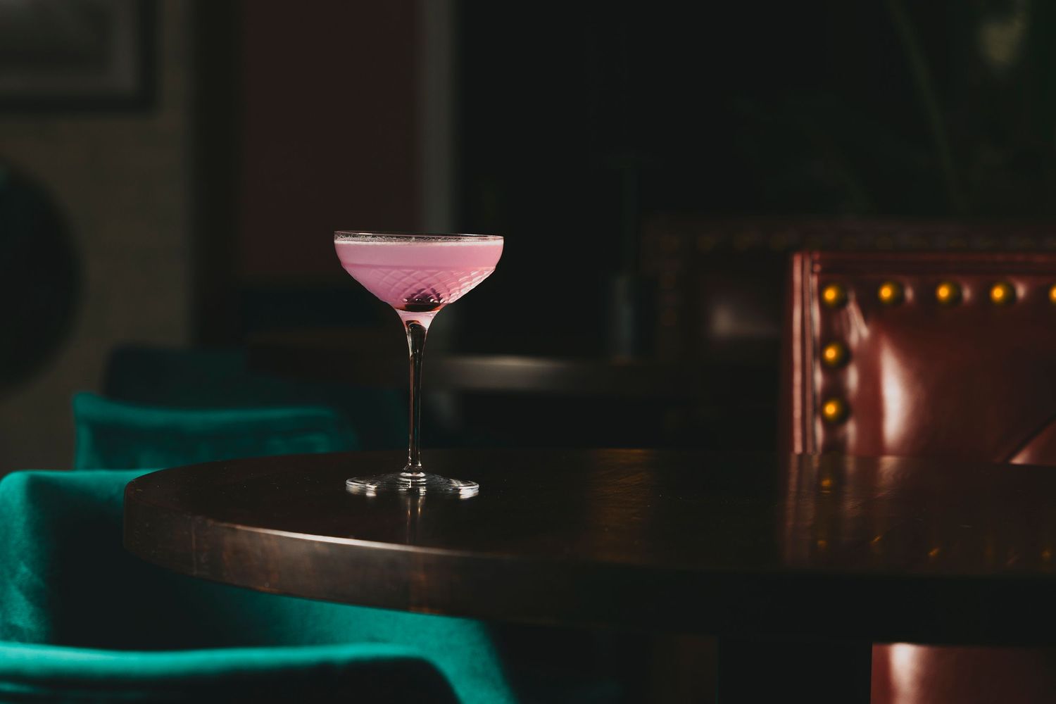 Pink cocktail in coupe glass on dark table with moody lighting and teal upholstery in background.