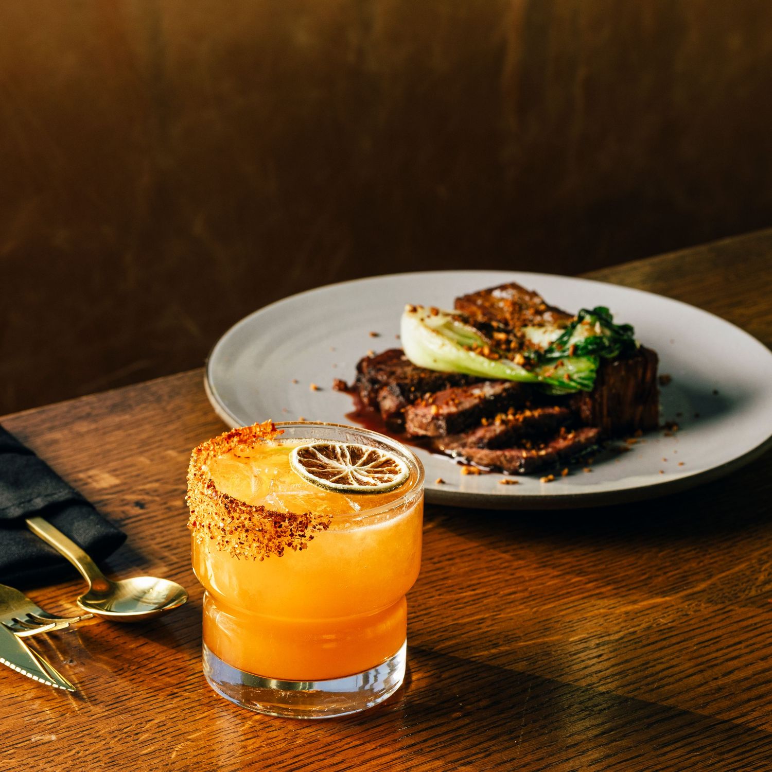 Orange cocktail garnished with lemon slice served alongside meat dish on wooden table.