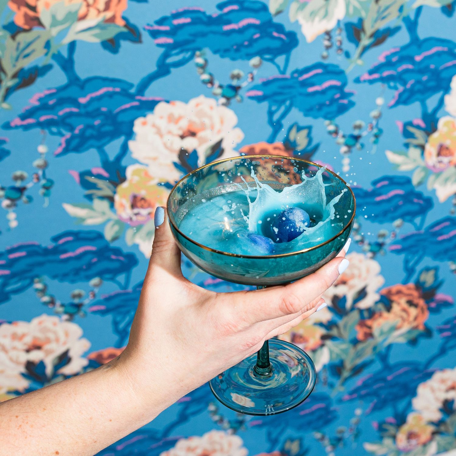 Hands holding turquoise cocktail glasses against blue floral wallpaper background.
