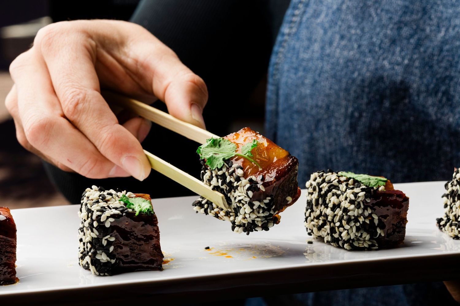 Hand using chopsticks to pick up sushi roll coated in black sesame seeds.