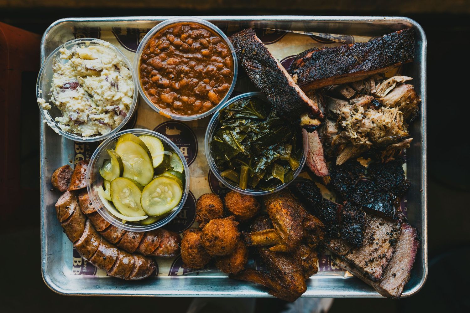 BBQ platter with brisket, ribs, pickles, beans, and sides served on a metal tray.