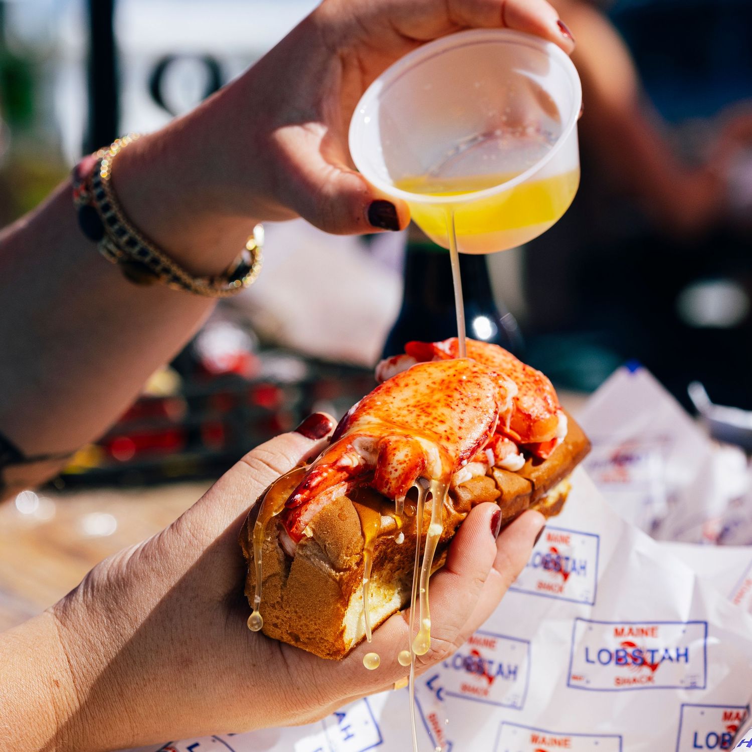 Hand holding a lobster roll while drizzling sauce, with beer glass in the background.
