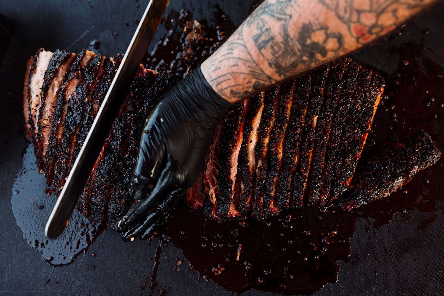 Gloved hands slicing smoked brisket on a black cutting board.