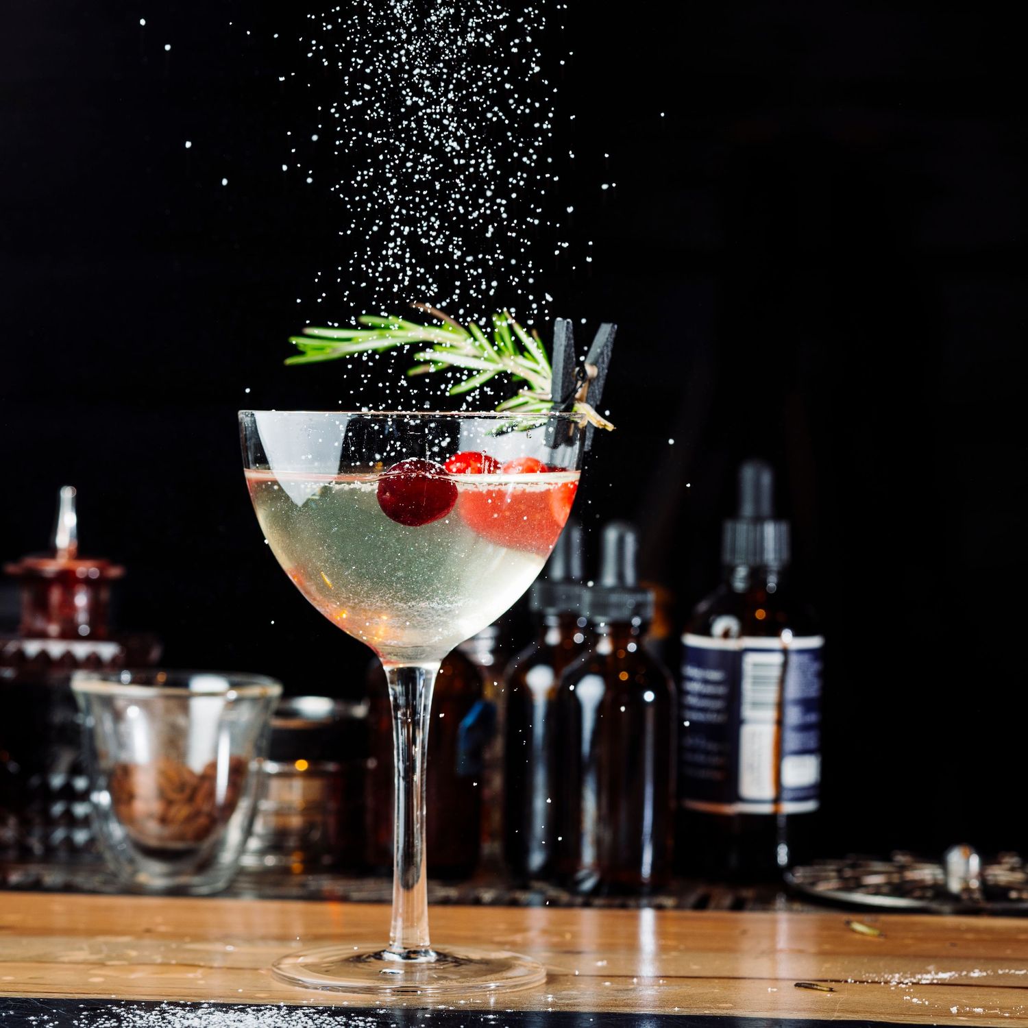 Elegant cocktail with raspberry and rosemary garnish being dusted with sugar against dark background.