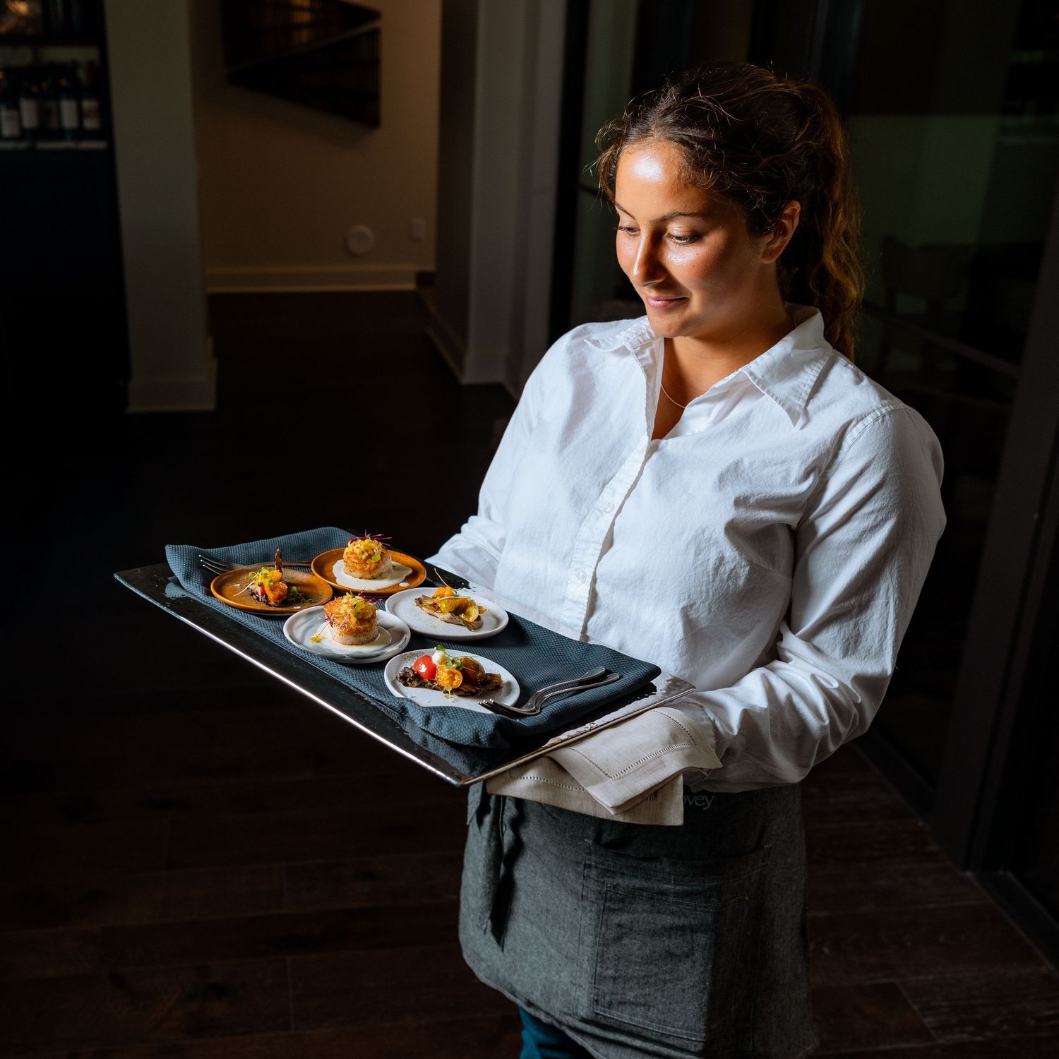 Server carrying multiple plates of food through a dimly lit restaurant dining room.