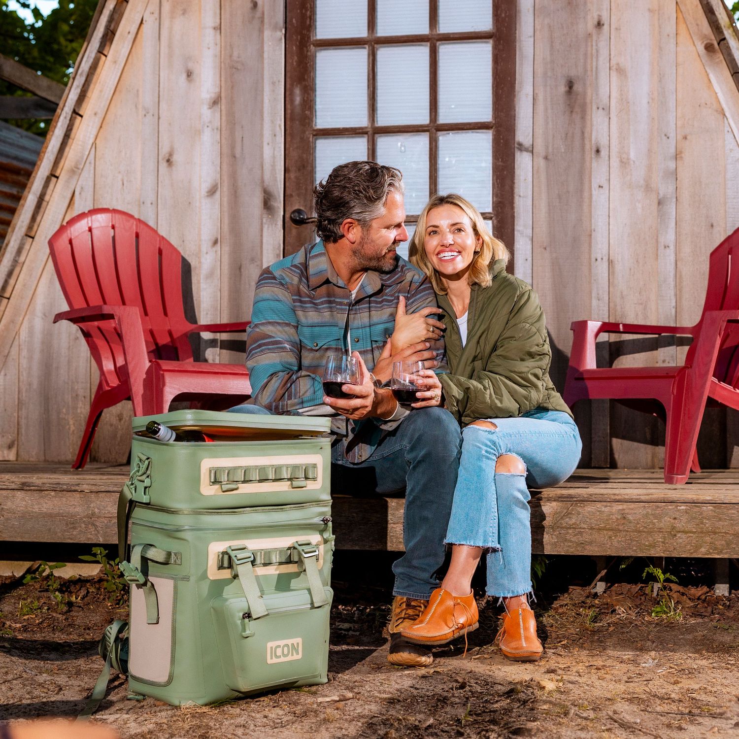 Friends relaxing with drinks outside rustic cabin with red Adirondack chairs.