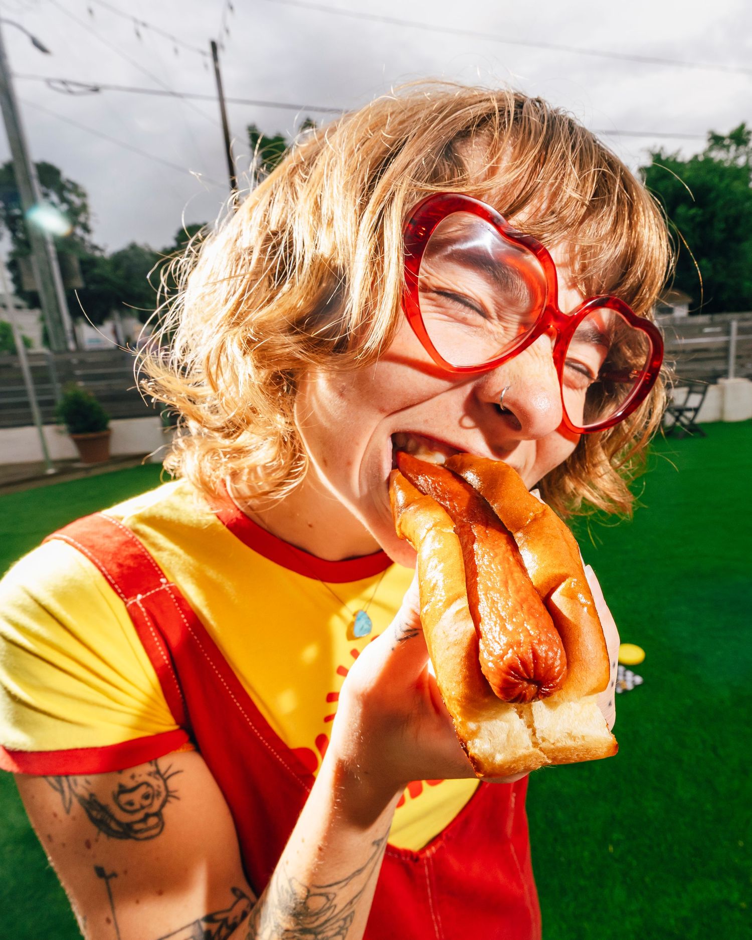 Person in a yellow shirt and red glasses takes an energetic bite of a sandwich outdoors on a sunny day.