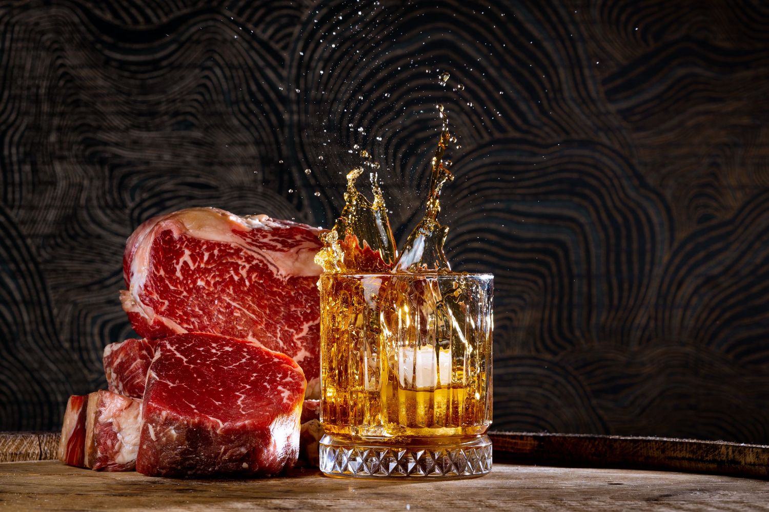 Raw steaks with whiskey glasses on dark textured background.