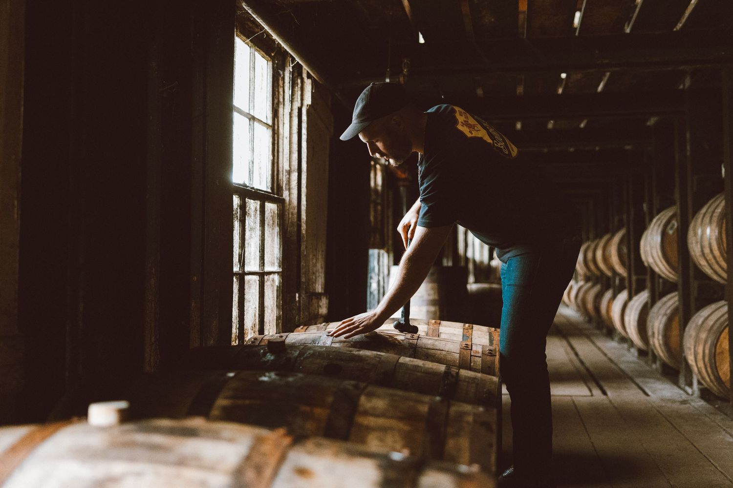 A silhouetted figure working in a dimly lit whiskey barrel warehouse with rows of wooden barrels stretching into the distance.