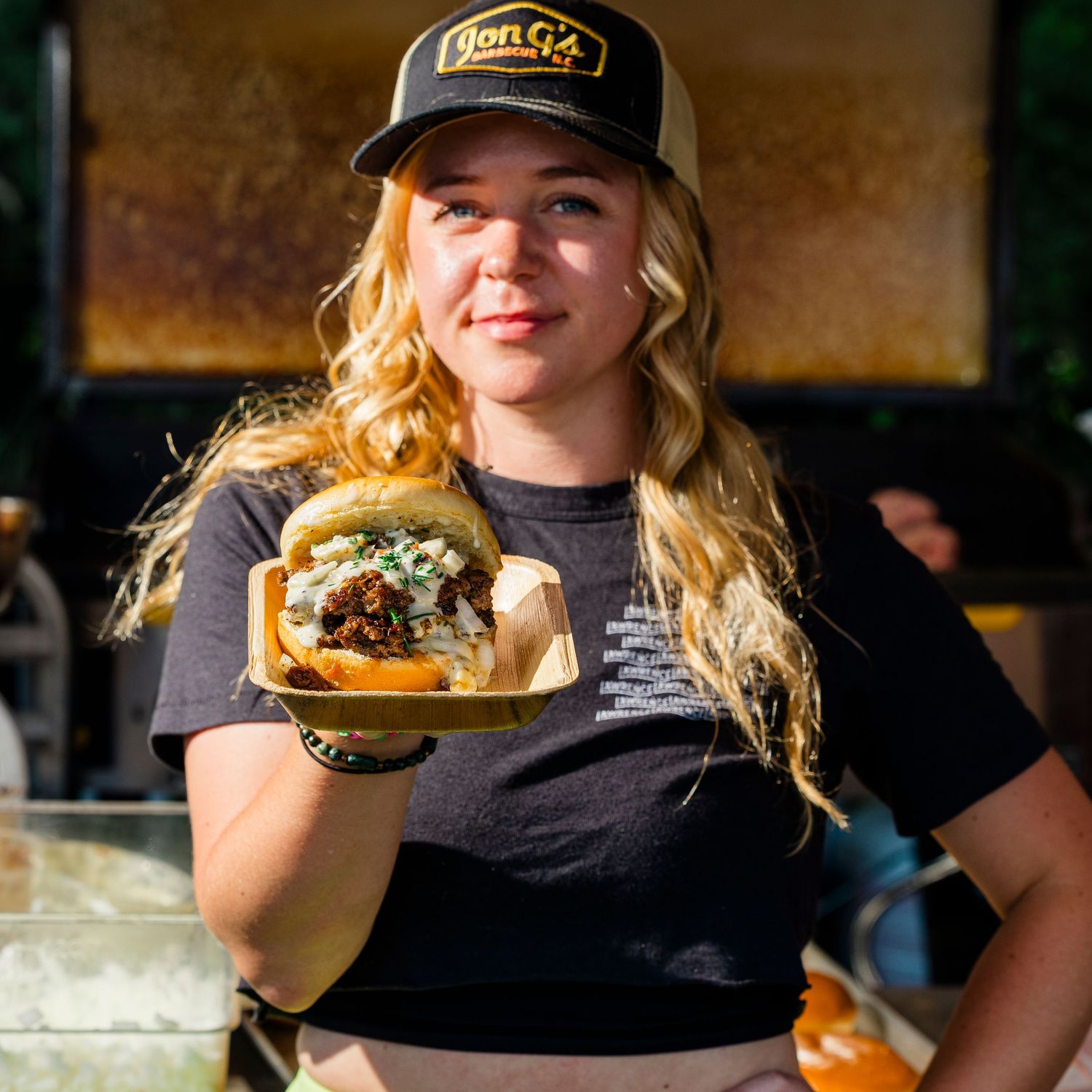 Restaurant server in black t-shirt and cap presents a gourmet burger on a wooden serving board.