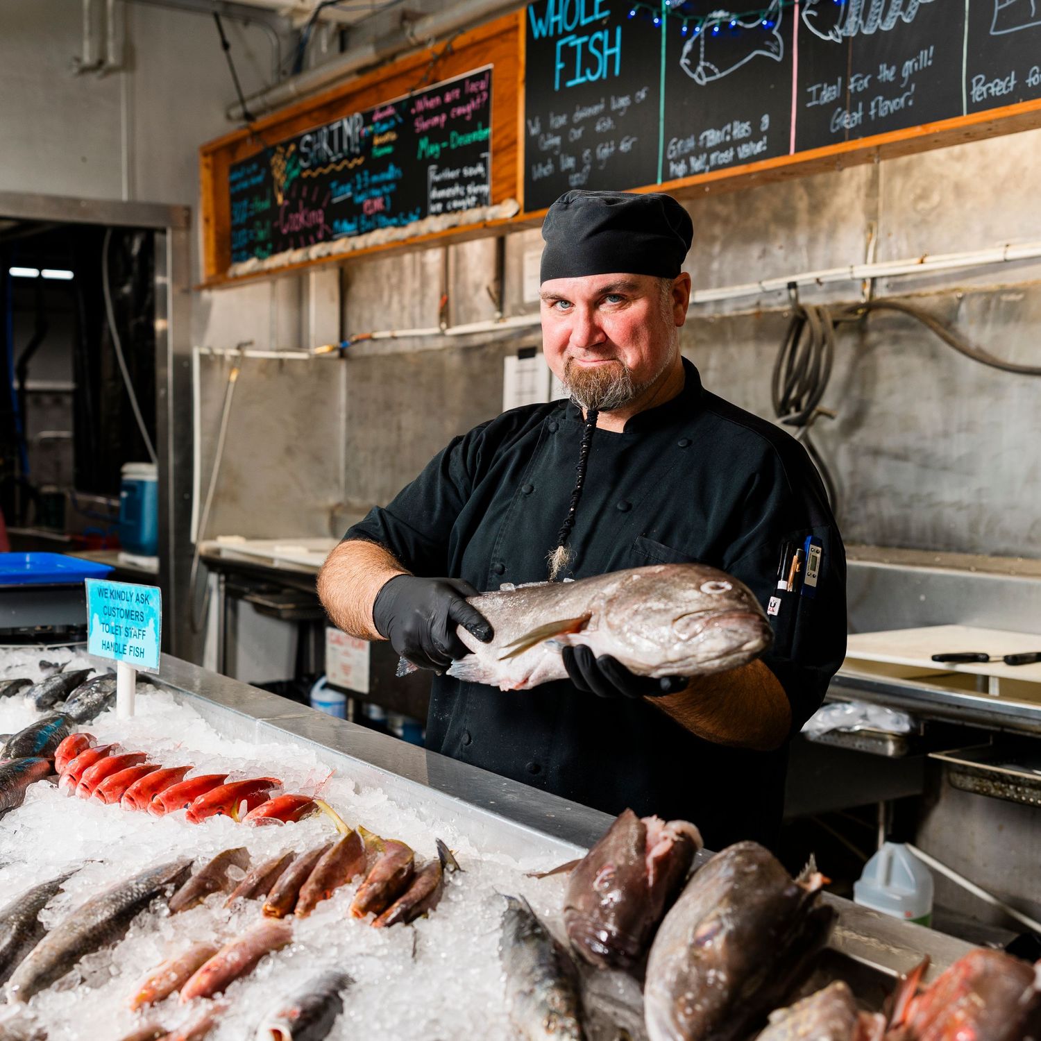A fishmonger in black attire preparing fresh fish at a seafood counter with a menu board above.