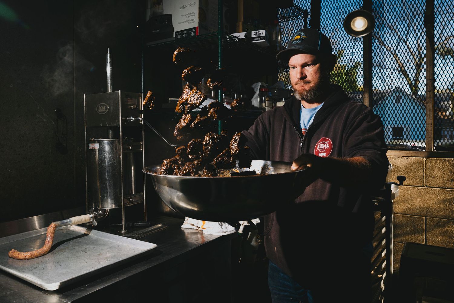 BBQ chef tending to a large pan of smoking meat on an outdoor cooker.