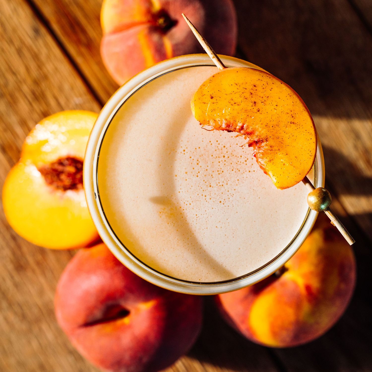 Fresh peach slices being added to a creamy cocktail in coupe glass.