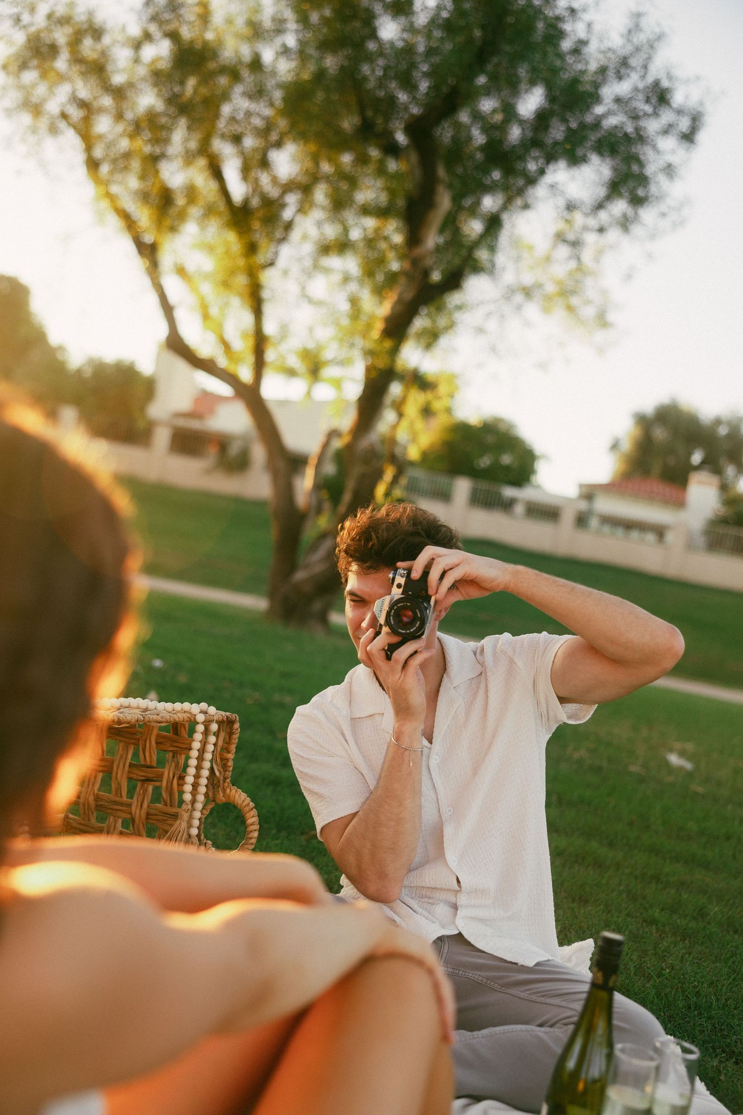 Person in white shirt taking photos with camera in park during golden hour sunset lighting.