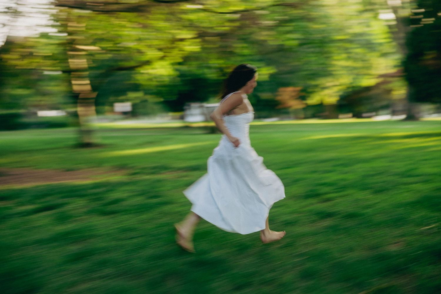 Motion-blurred image of someone in a flowing white dress running through a sunlit green park on a summer day.
