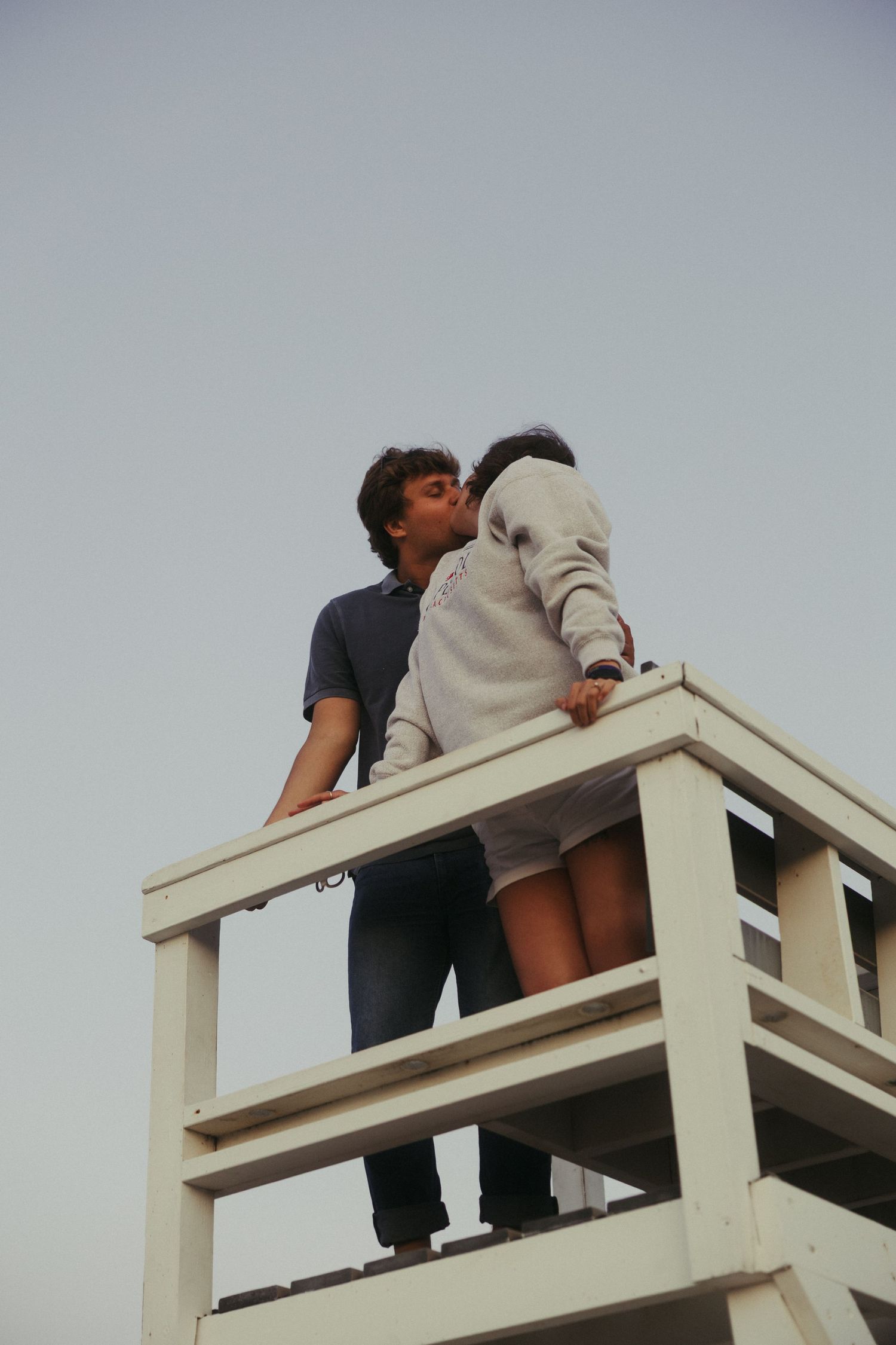 Two people sit playfully on a white wooden structure against an overcast sky.