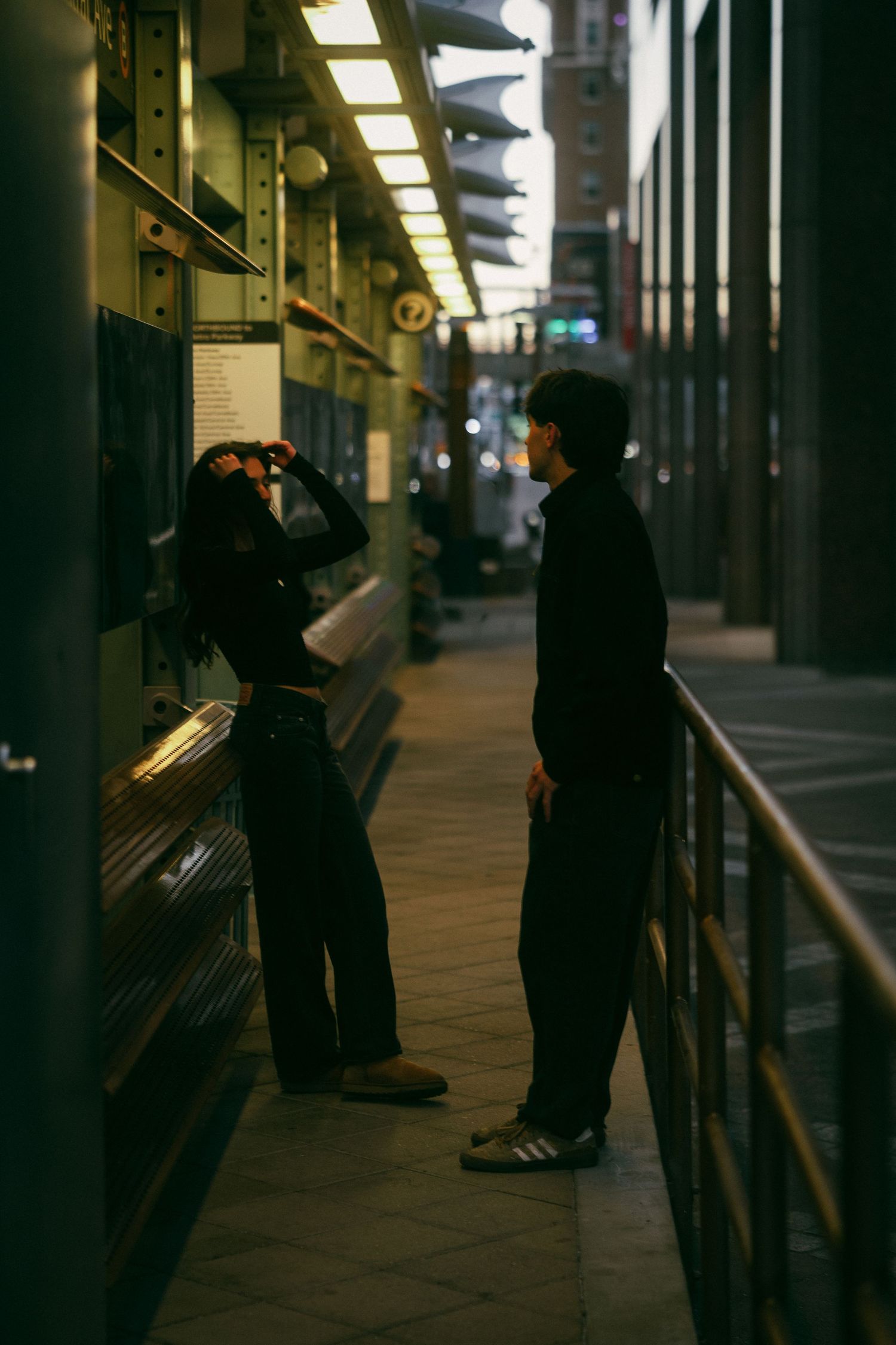Silhouettes of people waiting at a train station platform in low light conditions with fluorescent lighting overhead.