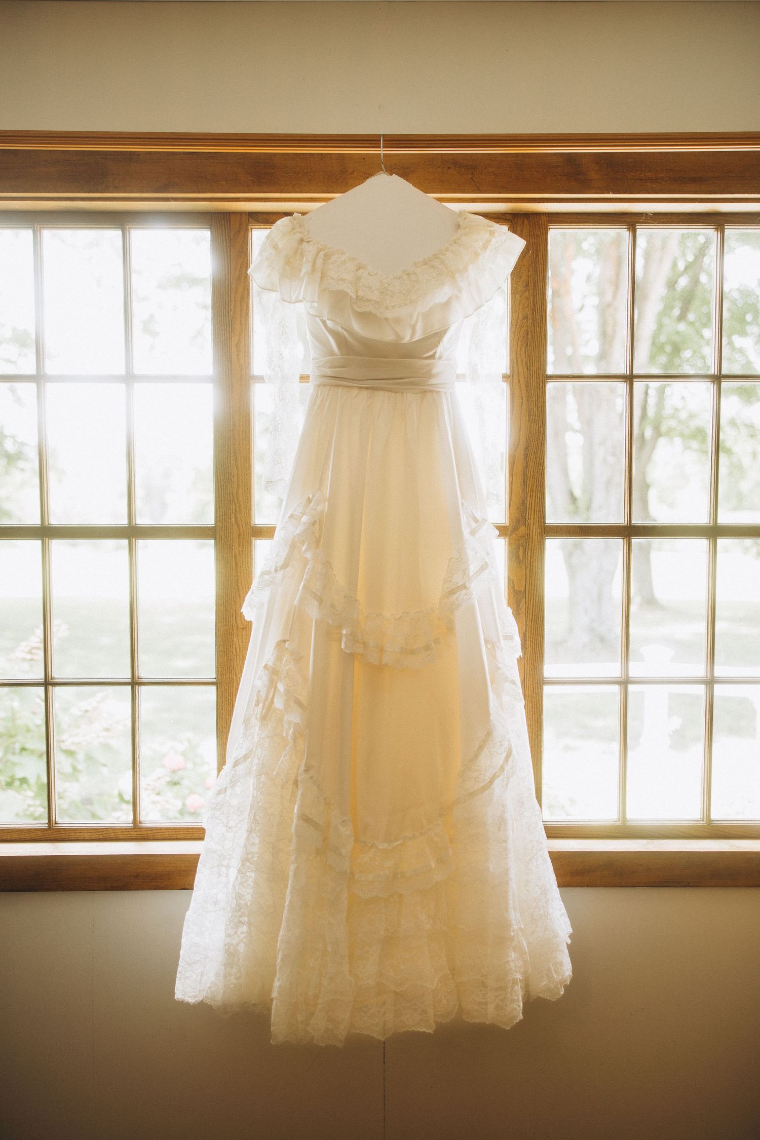 Vintage-style wedding dress hanging in window light with delicate lace details.