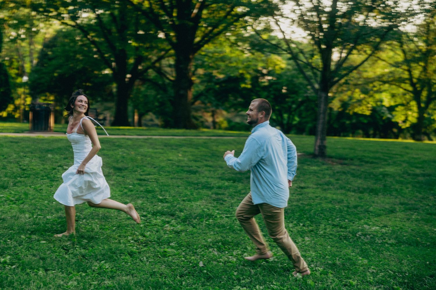 Couple playing and running through a sunlit park with trees in the background.