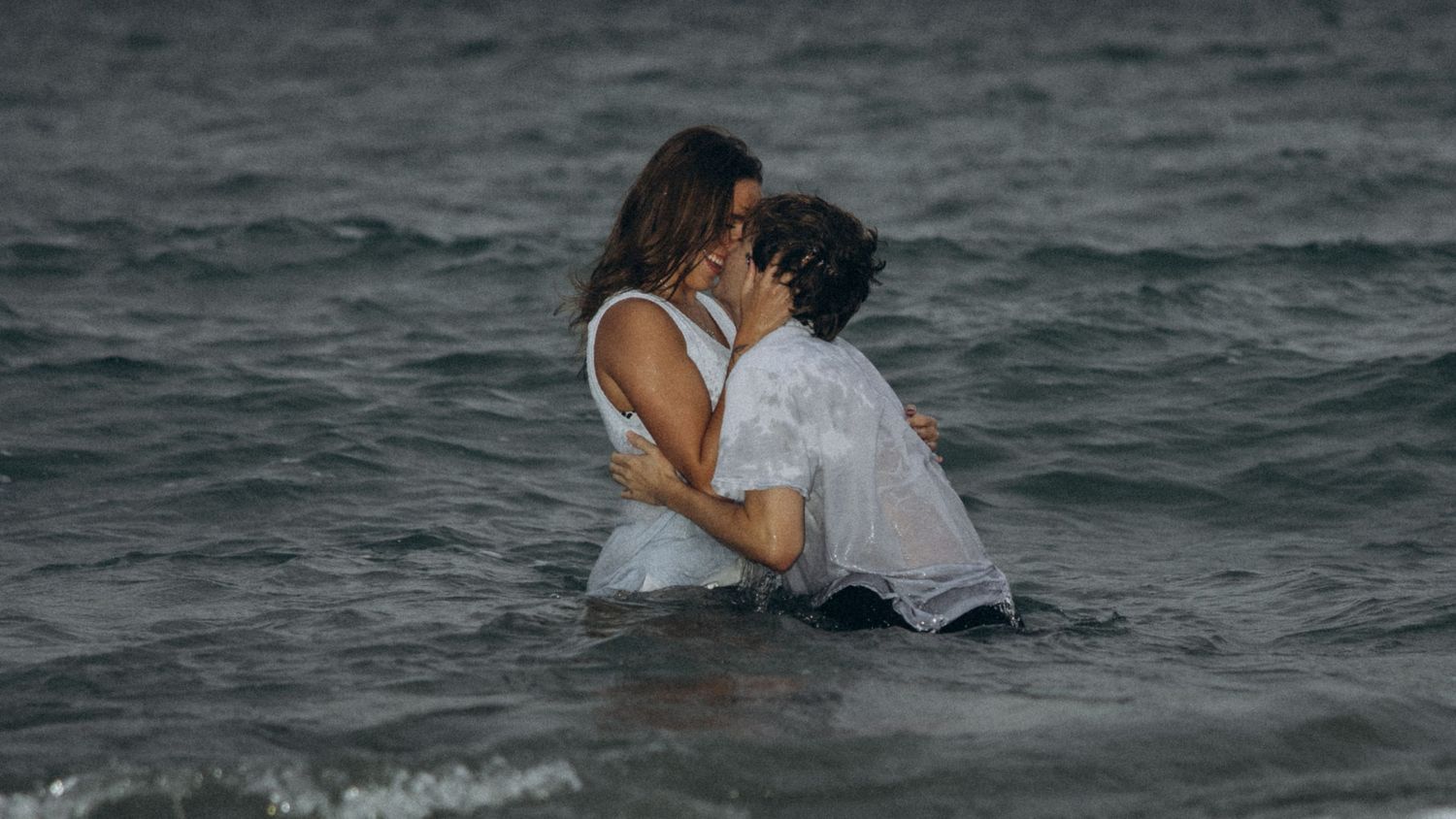 Romantic couple sharing a passionate kiss while standing in ocean waves, both wearing wet white clothing.