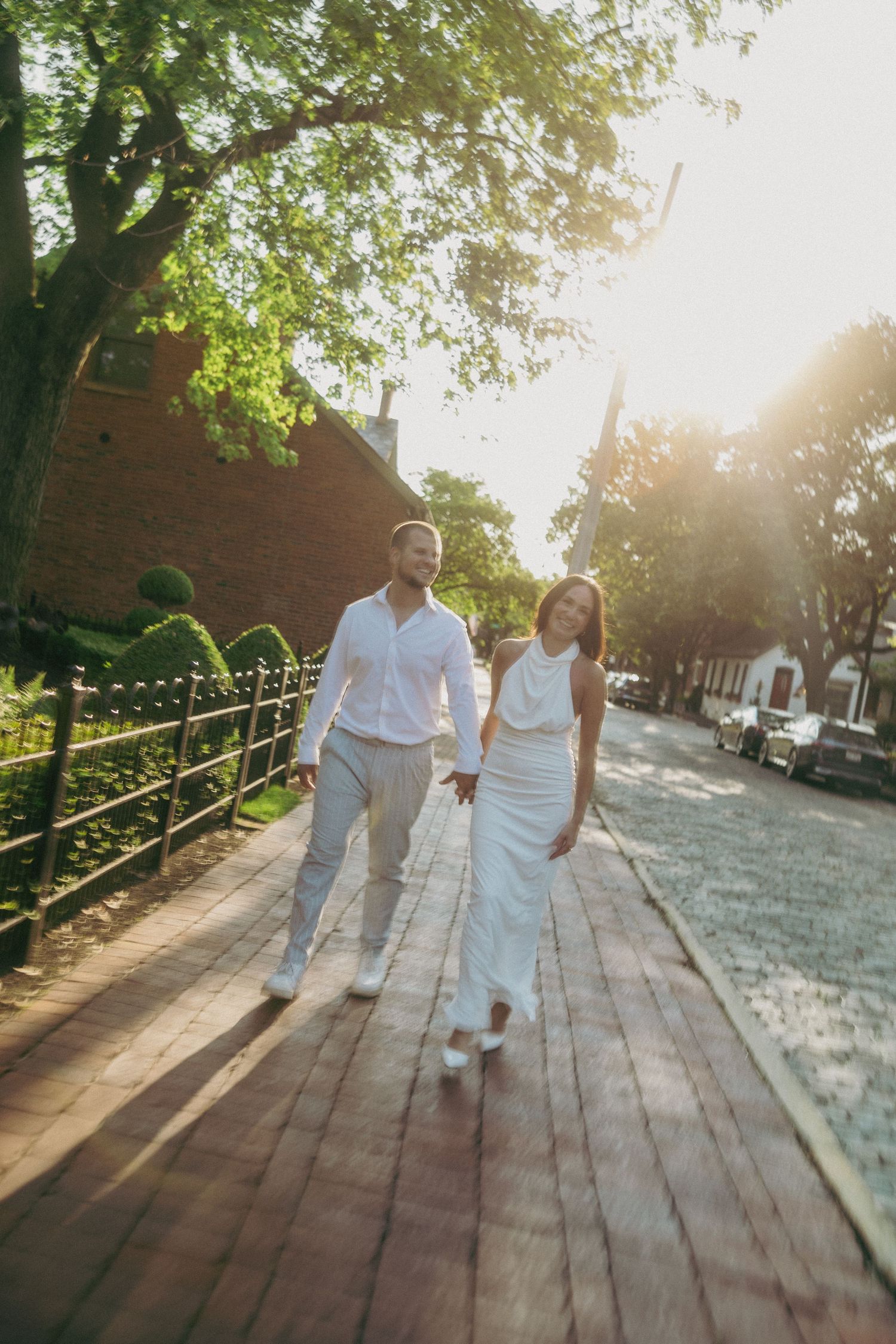 Couple in white outfits walking together on a wooden boardwalk during golden hour with sunlight streaming through trees.